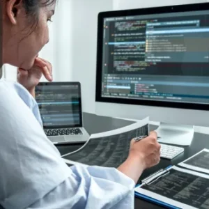 A woman analysing code on printed sheets while referencing programming scripts on a large desktop monitor and laptop screen in a modern workspace.