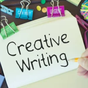 A hand writing "Creative Writing" in a notebook surrounded by colourful office supplies and clips.