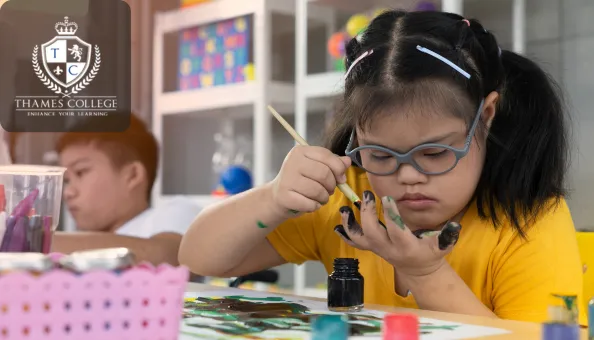 A young girl with autism focused on painting her hand with black paint while sitting at a table, with a boy in the background engaged in an activity.