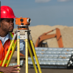 A building surveyor in a red hard hat and high-visibility vest using a theodolite on a construction site with heavy machinery in the background.
