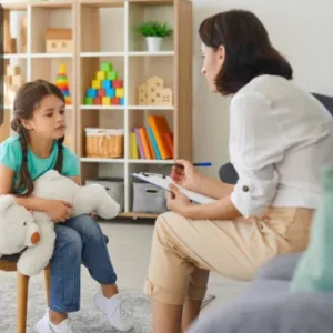 A child psychologist attentively talking with a young girl sitting on a chair with a teddy bear in a child-friendly room.