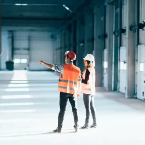 Two facilities professionals wearing high-visibility vests and hard hats inspect a large, empty industrial space with dock doors and overhead fixtures. The Thames College logo appears in the top-left corner.