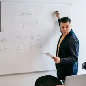 A male instructor solving fraction equations on a whiteboard during a Functional Maths class, holding notes and engaging with students in a classroom setting.