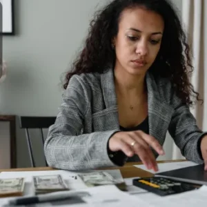 A focused woman working on a laptop with cash, payroll documents, and a calculator on the desk—illustrating payroll processing and financial records management.