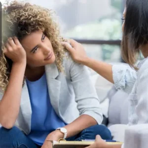 Two women engaged in a supportive conversation in a comfortable, light-filled room, one woman attentively listening and offering comfort to the other.