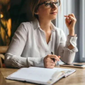 A professional woman sitting at a table with a pen and open notebook, appearing thoughtful while taking notes.