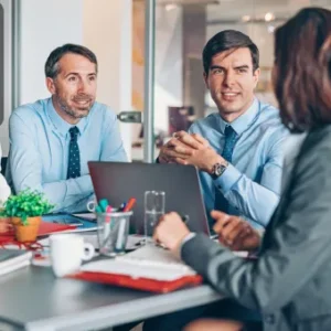 A group of office professionals in a meeting, discussing documents and working on a laptop at a conference table.