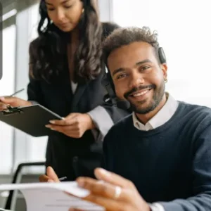 A smiling man wearing a headset sits at a desk with papers in hand, while a woman beside him takes notes on a clipboard. The Thames College logo is displayed in the top-left corner.