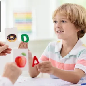 A young boy smiling and holding up alphabet flashcards during a phonics lesson at Thames College. A teacher’s hand is also visible holding flashcards with letters and pictures.