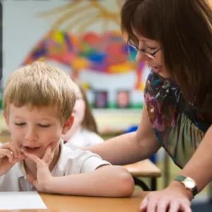 A primary school teacher supports a young boy as he works on an assignment in a colourful classroom setting. The Thames College logo is visible in the top left corner.