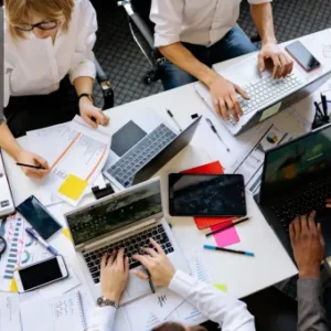 Team collaborating at a table filled with laptops, tablets, papers, and notes, representing an active project management workspace.