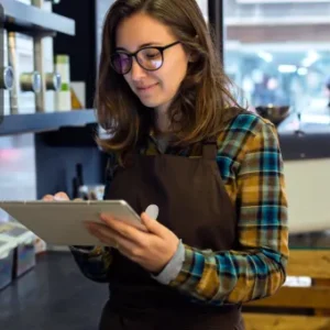 A retail worker in an apron uses a digital tablet behind the counter of a modern shop, surrounded by products and packaging materials. The Thames College logo appears in the top left corner.