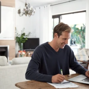 Smiling man working on a laptop and taking notes at a wooden desk in a bright, modern living room, with the Thames College logo in the corner.