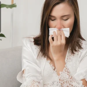 A woman sitting on a sofa using a tissue, showing symptoms of an allergic reaction.