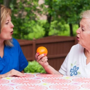 A speech therapist assisting an elderly woman with a speech exercise using an orange as a visual aid.