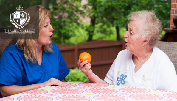 A speech therapist assisting an elderly woman with a speech exercise using an orange as a visual aid.