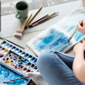 A person painting with watercolours in a sketchbook, surrounded by brushes, paints, and a cup of tea.