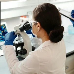 Scientist in a laboratory using a microscope for molecular biology research with colourful visuals displayed on a laptop screen.