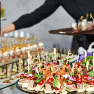 A catering professional arranges elegant appetisers on trays at a buffet table filled with colourful finger foods. The Thames College logo is visible in the corner.