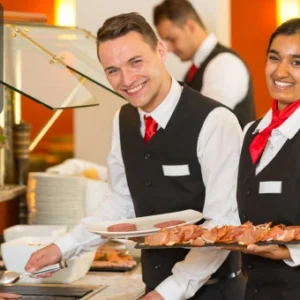 Two catering staff in uniform smiling while serving food at a buffet station in a restaurant setting. The Thames College logo is visible in the corner.