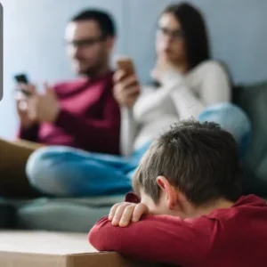 A young boy looking down with his head resting on his arms in the foreground, while his parents are distracted by their mobile phones on a sofa in the background.