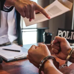 A handcuffed individual sits across a desk from a suited officer pointing at a legal document, with a “POLICE” sign visible in the background.