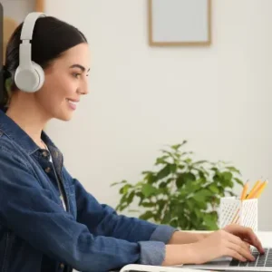 A woman wearing headphones smiles while working on a laptop at a bright, tidy desk, with plants and a notepad in the background. The Thames College logo is shown in the top left corner.