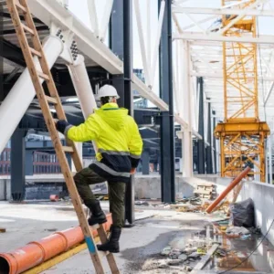Construction worker in high-visibility clothing climbing a wooden ladder at an active building site, surrounded by steel beams and cranes.