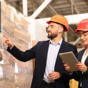 Two logistics managers wearing safety helmets inspecting stacked pallets in a warehouse, one pointing while the other uses a tablet.