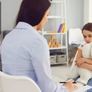 A child holding a teddy bear during a therapy session with a mental health professional taking notes.