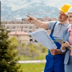 A property developer in a suit and construction worker wearing helmets examine building plans outdoors with a cityscape background.