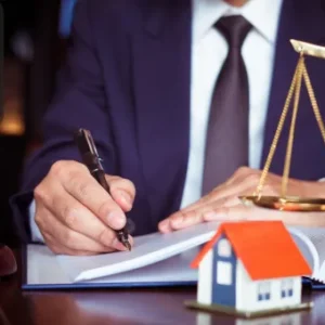 A professional in a suit signing legal documents with a small house model and scales of justice on the desk.