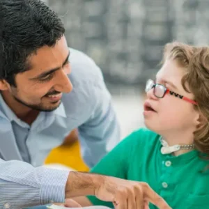 A smiling male teaching assistant supports a young student with special educational needs in a classroom setting. The child, wearing glasses and a green top, is engaged in conversation, while the Thames College logo is displayed in the top-left corner.