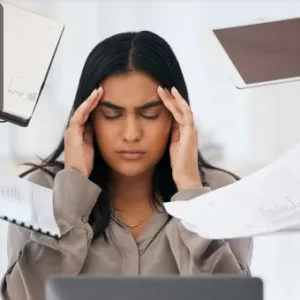 A stressed professional surrounded by documents, laptops, and tablets while holding her head in frustration.