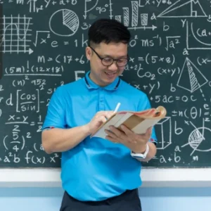 A teacher holding a book and smiling in front of a chalkboard filled with algebraic formulas and geometric diagrams.