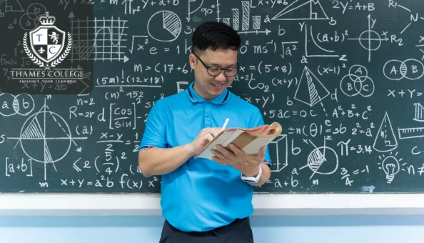 A teacher holding a book and smiling in front of a chalkboard filled with algebraic formulas and geometric diagrams.
