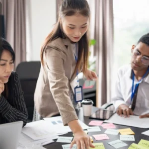Team of young professionals brainstorming around a table covered with colourful sticky notes and business documents, with the Thames College logo in the corner.