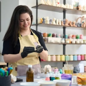 A smiling woman in an apron crafting candles in a well-organised workshop surrounded by colourful materials, wax, and fragrance oils, with the Thames College logo in the top-left corner.
