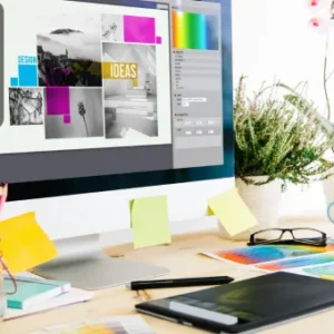 A modern graphic designer’s workspace with a desktop monitor displaying a collage design project and colour palette tools, surrounded by creative supplies.