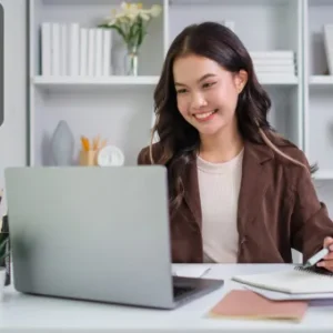 A smiling young woman working on a laptop at a neatly organised desk, taking notes in a notebook during an online session.