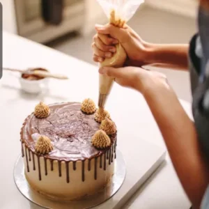 A person decorating a chocolate-drizzled cake with piped frosting swirls using a piping bag.