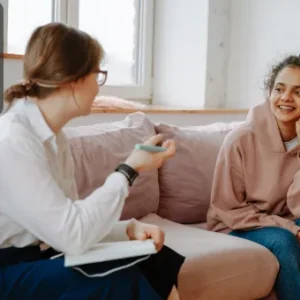 A counsellor speaking with a smiling young woman in a comfortable setting, both seated on a sofa during a session.