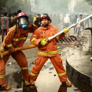 Firefighters in full protective gear, including helmets and masks, using a high-pressure hose to control a fire in an urban area, with the Thames College logo in the top left corner.
