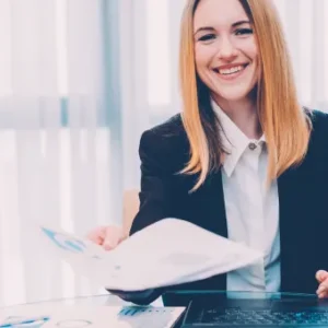 Smiling businesswoman handing over a document across a desk with a laptop and reports.