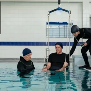 Two hydrotherapy practitioners assist a patient being lowered into a therapeutic pool using a hoist, with the Thames College logo in the top left corner.