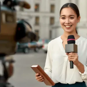 Smiling female journalist holding a microphone and notebook during an outdoor news report.