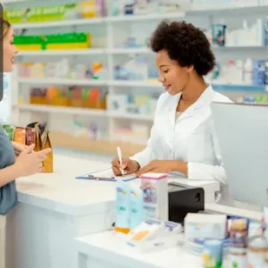 A pharmacist in a white coat assisting a customer at a pharmacy counter, surrounded by shelves of medicines and health products.