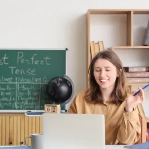 Smiling young woman teaching English grammar online with a laptop, books, and a blackboard showing the past perfect tense.