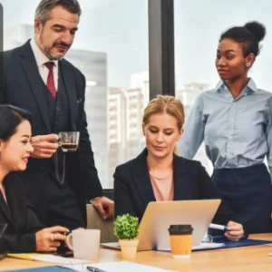 Group of diverse business professionals collaborating around a table with a laptop and documents in an office setting, with the Thames College logo in the corner.