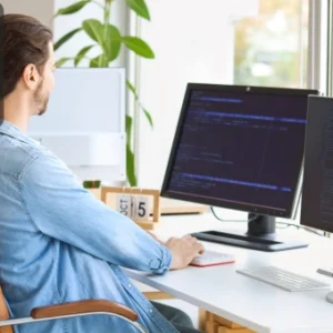 Man sitting at a desk working on dual computer monitors displaying code and data, with the Thames College logo in the corner.